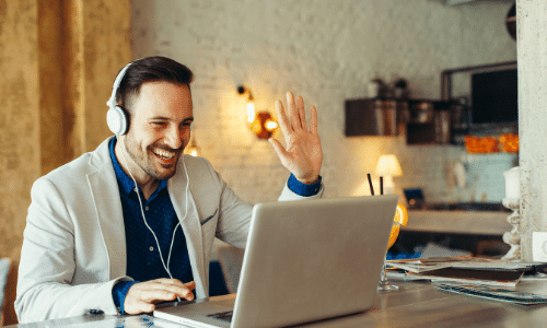 man is wearing headphones and attending to a virtual meeting on his laptop while smiling and raising his left hand