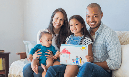 a multiracial family is sitting on a couch and smiling with a white mother, a black father, a daughter holding a picture that she drew of her family with crayons, and a baby boy