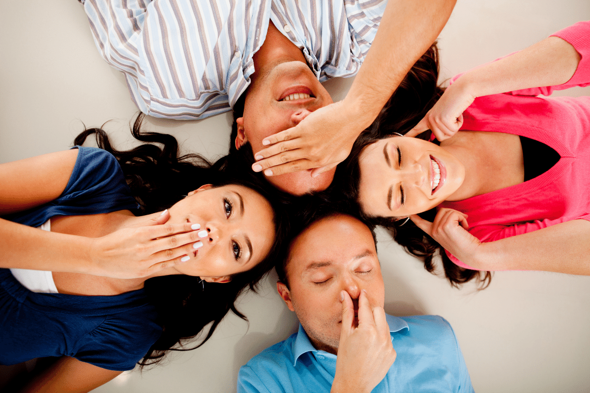 4 people lying down: one man is covering eyes with his right hand, a woman is plugging her ears with both hands, a man is pinching his nose, and a woman is covering her mouth with her right hand