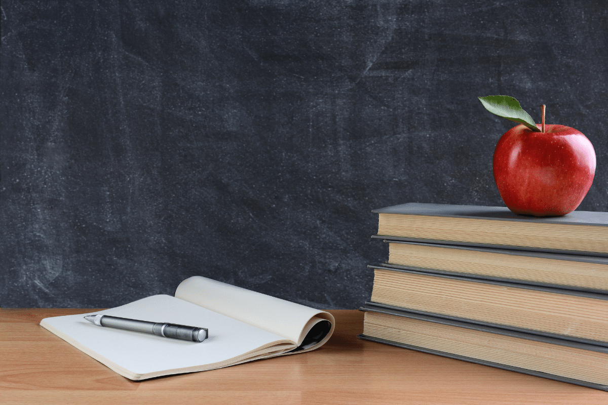 teacher's desk sits in front of a chalkboard; on the left side of the desk is an open notebook with a black pen on top; on the right side of the desk sits a stack of 4 gray books with a red apple on top