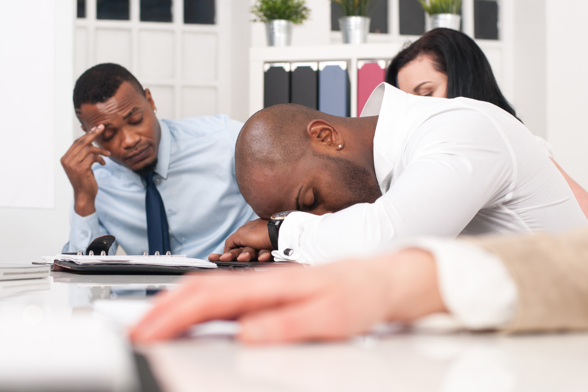 3 bored adults in a training- african american man wearing white shirt is sleeping on his hands, brunette woman has her eyes closed, african american man with blue short has right hand on his eyebrow as if he has a headache
