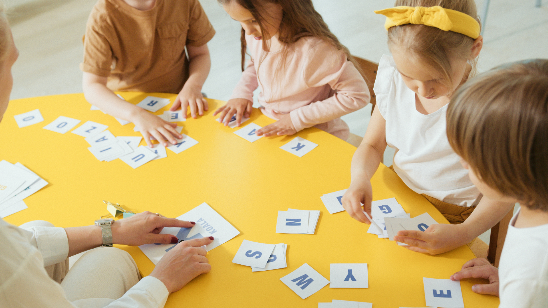 One teacher and four young students sit at a yellow table and move letter tiles around to spell words.